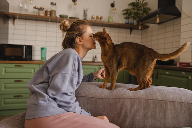 Young caucasian blonde woman with casual clothes is in contact with cat while sitting on couch pet concept | The Web Univers