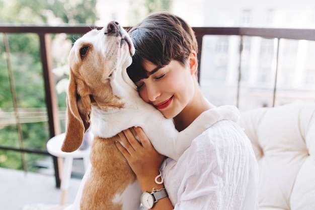 Close-up portrait of pleased woman with short brown hair embracing funny beagle dog with eyes closed | The Web Univers