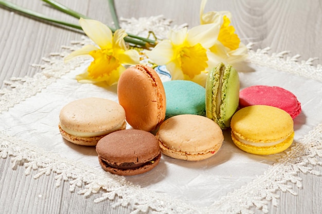 Sweet macarons cakes of different color on silk napkin with bouquet of fresh yellow daffodils on wooden background. | The Web Univers